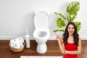 Tazza del WC con cubetti di ghiaccio, di lato scopino, rotoli di carta igienica. Dall'altro lato pianta e donna sorridente che indica il WC